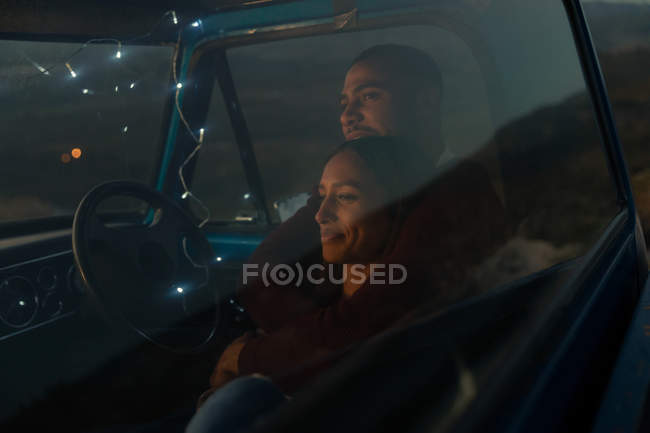 Vista lateral de una joven pareja mixta sentada en su camioneta, sonriendo y abrazándose al atardecer durante una parada en un viaje por carretera . - foto de stock