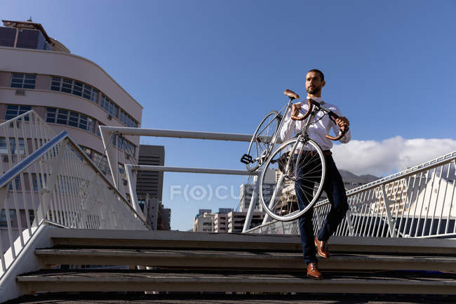Vista frontal do jovem caucasiano carregando sua bicicleta para baixo degraus na cidade. Digital Nomad em movimento . — Fotografia de Stock