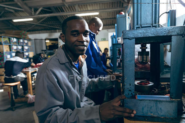 Portrait close up of a young mixed race man in sports clothing factory, looking to camera and smiling with coworkers working in the background. — Stock Photo