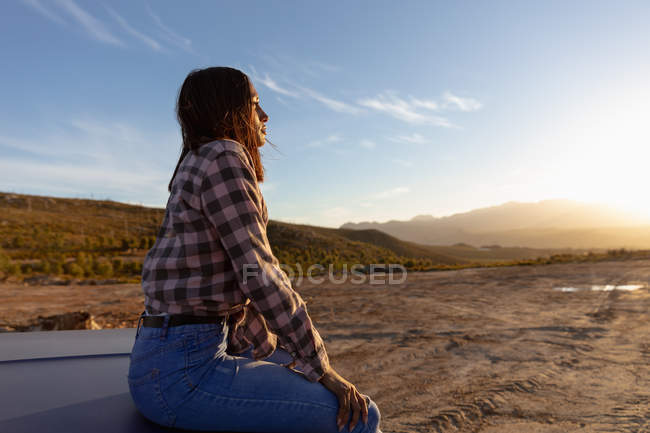 Vista laterale ravvicinata di una giovane donna seduta sul cofano di un pick-up che si gode la vista al tramonto durante una sosta durante un viaggio in auto — Foto stock