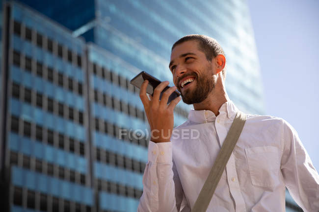 Vista frontal de perto de um jovem caucasiano sorridente falando em um smartphone segurando-o na frente de seu rosto em uma rua da cidade. Digital Nomad em movimento . — Fotografia de Stock
