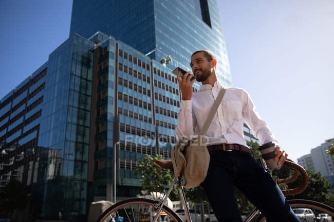 Vista frontal de perto de um jovem caucasiano segurando um café takeaway e falando em um smartphone segurando-o na frente de seu rosto, enquanto se inclina em sua bicicleta em uma rua da cidade. Digital Nomad em movimento . — Fotografia de Stock