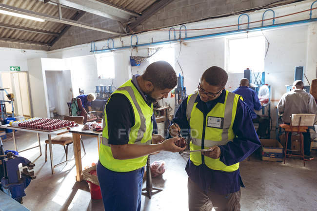 Vue de face d'un jeune manager masculin mixte portant des lunettes tenant un ordinateur tablette et parlant avec un travailleur masculin mixte et inspectant une balle qu'il tient, dans une usine de ballon de cricket . — Photo de stock