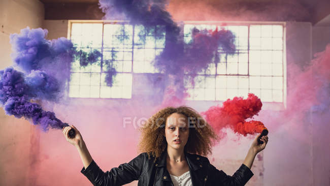 Vorderansicht einer jungen kaukasischen Frau mit lockigem Haar, die eine Lederjacke trägt, während sie aufmerksam in die Kamera blickt und eine Rauchmaschine in der Hand hält, die roten und blauen Rauch in einer leeren Lagerhalle produziert — Stockfoto