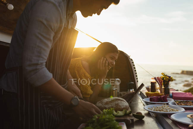 Вид сбоку на молодую смешанную расовую пару, занятую приготовлением пищи, стоя в фургоне с открытым верхом, предлагающем широкий ассортимент еды на продажу, женщина держит в руках планшетный компьютер, они освещаются солнечным светом — стоковое фото