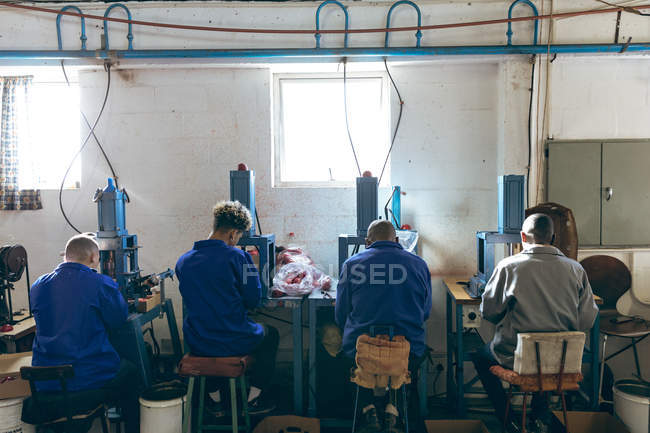 Rear view of a diverse group of four male workers sitting and operating machines at a factory making cricket balls. — Stock Photo