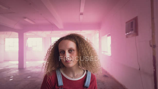 Vue de face d'une jeune femme caucasienne aux cheveux bouclés regardant attentivement la caméra tout en se tenant à l'intérieur d'un entrepôt vide avec du brouillard rose — Photo de stock