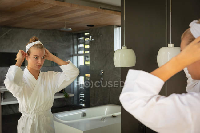 Primer plano de una joven mujer caucásica vestida con una bata de baño mirándose en el espejo y preparando su cabello en un baño moderno . - foto de stock
