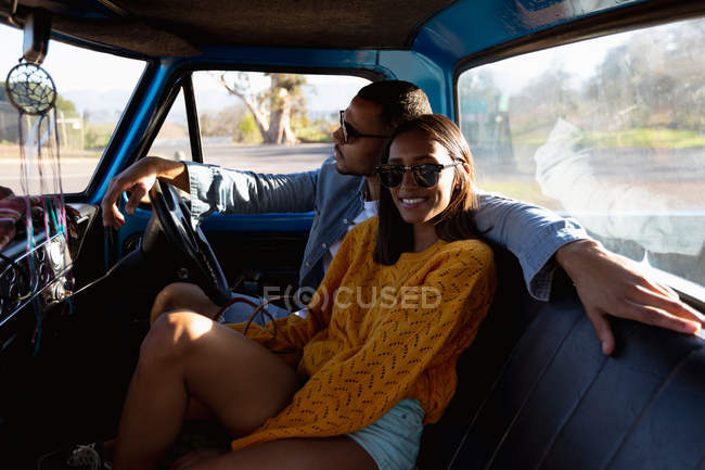 Vista laterale di una giovane coppia mista seduta nel loro pick-up, sorridente e abbracciata durante un viaggio su strada. L'uomo sta guidando e la donna si gira verso la telecamera e sorride — Foto stock