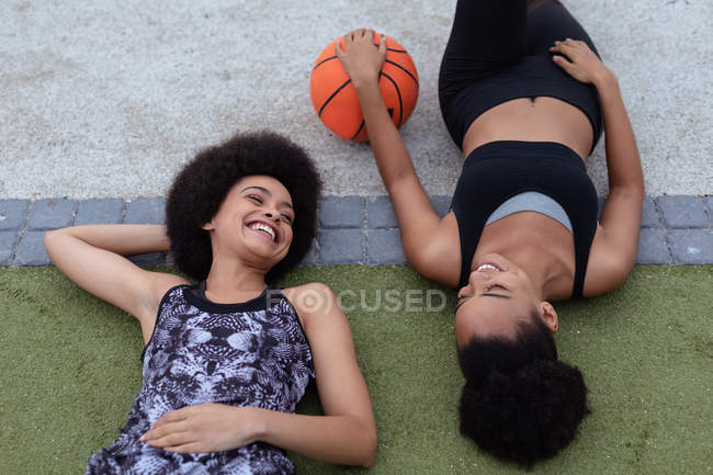 Elevado de cerca de dos hermanas adultas jóvenes de raza mixta con ropa deportiva, tumbadas en el suelo, mirándose y sonriendo, una sosteniendo una pelota de baloncesto - foto de stock