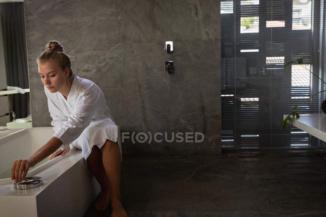 Вид спереди на молодую кавказскую женщину, текущую воду и сидящую на ванне в халате в современной ванной комнате . — стоковое фото