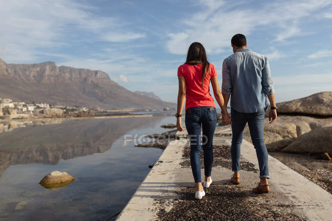 Visão traseira close-up de um jovem casal de raças mistas de mãos dadas andando em um molhe junto ao mar, com céu azul e montanhas no fundo — Fotografia de Stock
