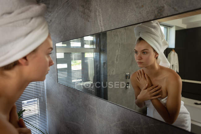 Vista laterale da vicino di una giovane donna caucasica che indossa un asciugamano da bagno e con i capelli avvolti in un asciugamano, guardando allo specchio in un bagno moderno
. — Foto stock