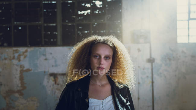 Frontansicht einer jungen kaukasischen Frau mit lockigem Haar, die eine Lederjacke trägt, während sie in einer leeren Lagerhalle mit Sonnenlicht in die Kamera blickt — Stockfoto