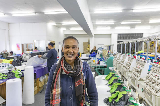 Ritratto da vicino di una giovane donna di razza mista che indossa una sciarpa, in piedi accanto a una fila di macchine in una fabbrica di abbigliamento sportivo illuminata, guardando la fotocamera e sorridendo . — Foto stock