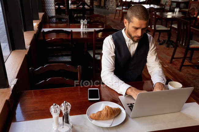 Elevated view of a young Caucasian man using a laptop computer sitting at a table inside a cafe. Digital Nomad on the go. — Stock Photo
