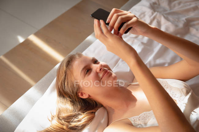 Primer plano de una joven rubia caucásica sonriente acostada boca arriba en la cama usando un smartphone . - foto de stock