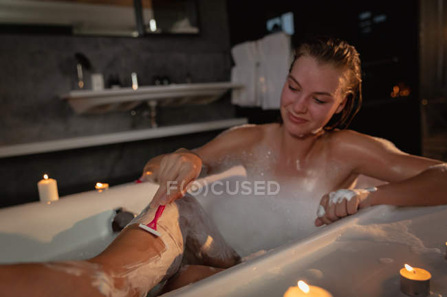 Front view of smiling young Caucasian woman sitting in the bath shaving her legs, with lit candles around the bathtub. — Stock Photo