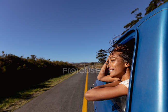 Vue latérale d'une jeune femme de race mixte penchée par la fenêtre avant côté passager d'une camionnette souriante, alors qu'elle descend l'autoroute lors d'un voyage en voiture — Photo de stock
