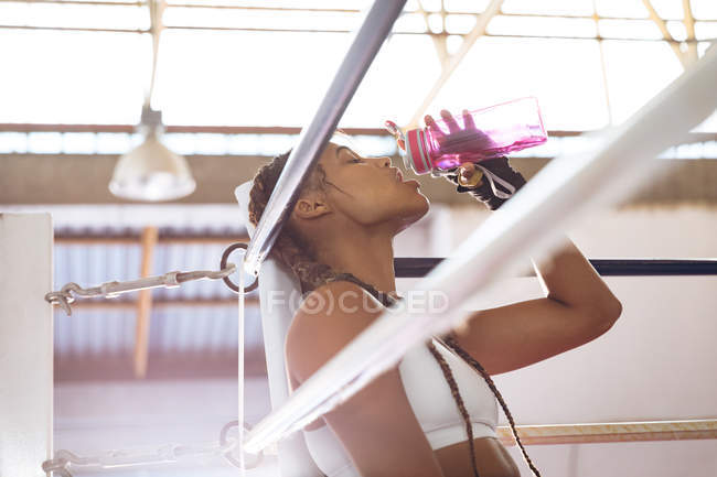 Vista lateral de boxeador fêmea bebendo água no ringue de boxe no centro de fitness. Forte lutador feminino no treinamento de ginásio de boxe duro . — Fotografia de Stock