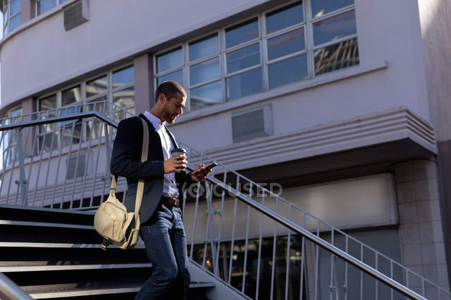 Seitenansicht eines jungen kaukasischen Mannes, der eine Umhängetasche trägt, sein Smartphone benutzt und einen Kaffee zum Mitnehmen in der Hand hält, während er die Treppe nach draußen geht. Digitaler Nomade unterwegs. — Stockfoto