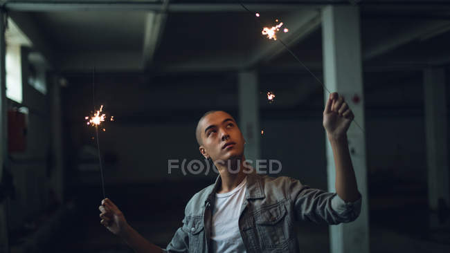 Frontansicht eines jungen hispanisch-amerikanischen Mannes, der eine graue Jacke über einem weißen Hemd trägt und von der Kamera wegschaut, während er in einem leeren Lagerhaus an beiden Händen brennende Funken hält — Stockfoto