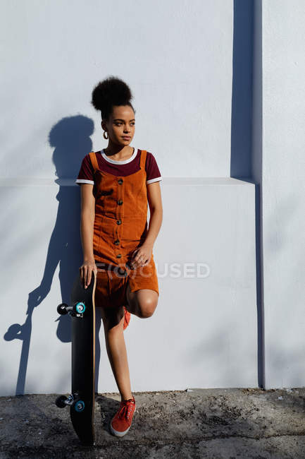 Frontansicht einer jungen Frau in einem Kleid, die in einer städtischen Straße in der Sonne an eine Wand gelehnt ist und ein Skateboard hält — Stockfoto