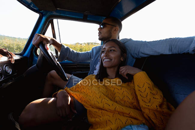 Vue latérale rapprochée d'un jeune couple de course mixte assis dans leur pick-up lors d'un voyage en voiture. L'homme conduit et la femme s'appuie sur lui et sourit — Photo de stock