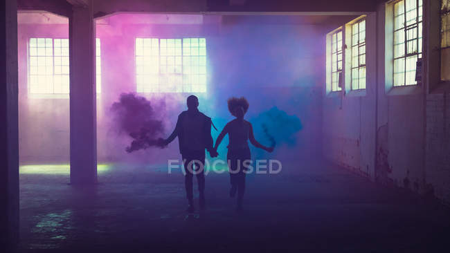 Vista frontal de la silueta de una pareja con fabricantes de humo que producen humo violeta y azul mientras se toman de la mano y corren dentro de un almacén vacío - foto de stock