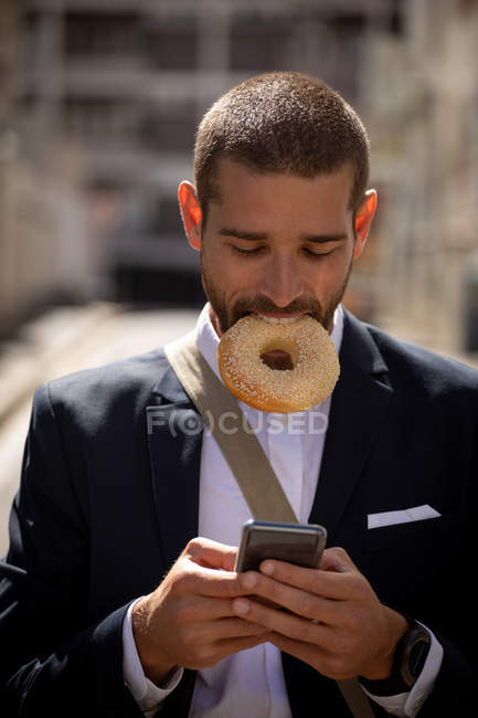 Вид спереду крупним планом молодого Кавказького чоловіка тримає кільцевий пончик в роті і використовуючи свій смартфон на міській вулиці. Цифрові кочівники на ходу. — стокове фото