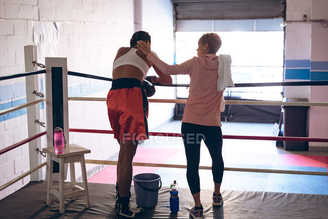 Задний вид тренера, утешающего женщину-боксера в боксерском ринге в фитнес-центре. Сильная женщина-боец в боксёрском зале тяжело тренируется
. — стоковое фото