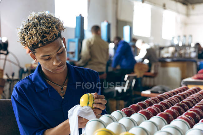Вид спереди на молодого смешанного гонщика, сидящего полирующего крикетные мячи в конце производственной линии на заводе по производству крикетных мячей. На заднем плане коллеги работают с машинами на рабочих станках . — стоковое фото