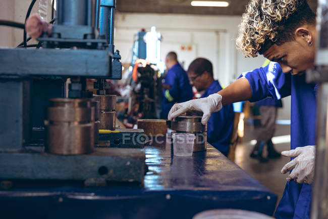 Вид сбоку на молодого смешанного гонщика в перчатках и комбинезоне, готовящего смесь для производственной линии на заводе по производству мяча для крикета, с коллегами, работающими машинами на заднем плане . — стоковое фото
