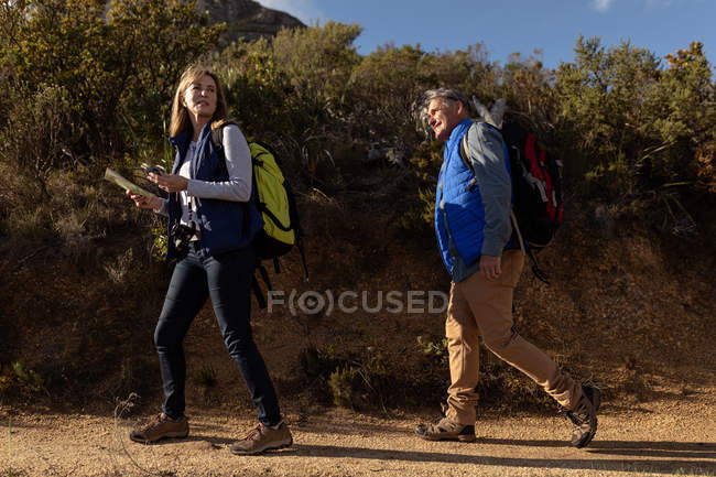 Side view close up of a mature Caucasian woman and man wearing backpacks walking single file along a trail during a hike in the countryside, the woman is holding a map and a compass — Stock Photo