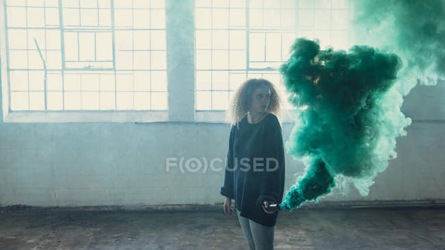 Vista lateral de una joven mujer caucásica con el pelo rizado usando una manga larga negra y sosteniendo una máquina de humo produciendo humo verde dentro de un almacén vacío - foto de stock