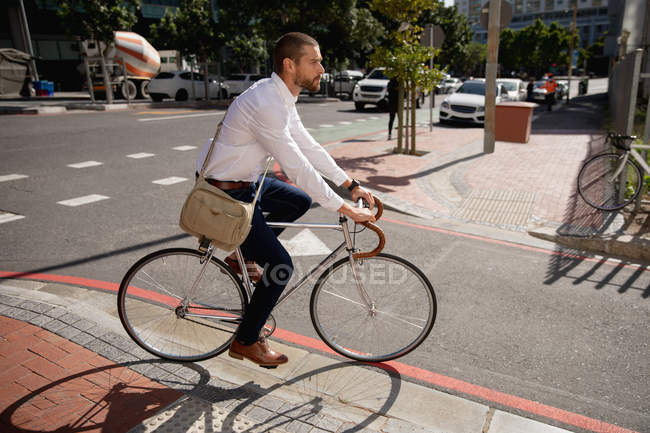 Side view of a young Caucasian man carrying a shoulder bag riding his bicycle on a road in the city. Digital Nomad on the go. — Stock Photo