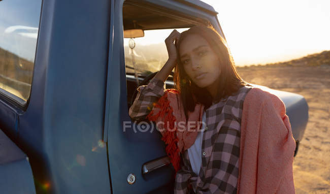 Portrait d'une jeune femme métissée réfléchie avec une couverture sur les épaules, penchée sur la porte d'une camionnette regardant droit vers la caméra au coucher du soleil, lors d'un arrêt sur une route — Photo de stock