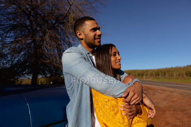 Vista lateral de una joven pareja mixta apoyada en su camioneta, abrazando y disfrutando de la vista durante una parada en un viaje por carretera - foto de stock