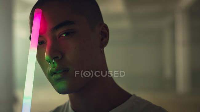 Close up view of a young Hispanic-American man with piercing on both ears and nose wearing a white sleeveless holding a light stick looking intently at the camera — Stock Photo