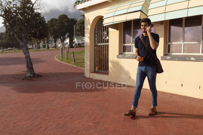 Вид спереди улыбающегося молодого смешанного гонщика, несущего плечевую сумку, держащего кофе на вынос и говорящего по смартфону, выходящего из здания на солнце — стоковое фото