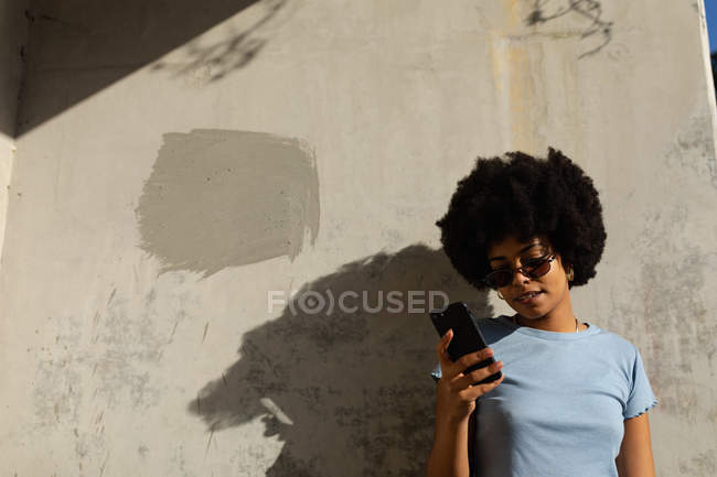 Vista frontal close-up de uma jovem mulher de raça mista usando óculos de sol em pé contra uma parede usando seu smartphone ao sol — Fotografia de Stock