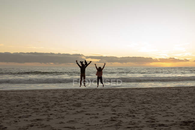 Задний вид зрелого кавказца, прыгающего вместе с руками в воздухе на пляже, обращенном к морю на закате — стоковое фото