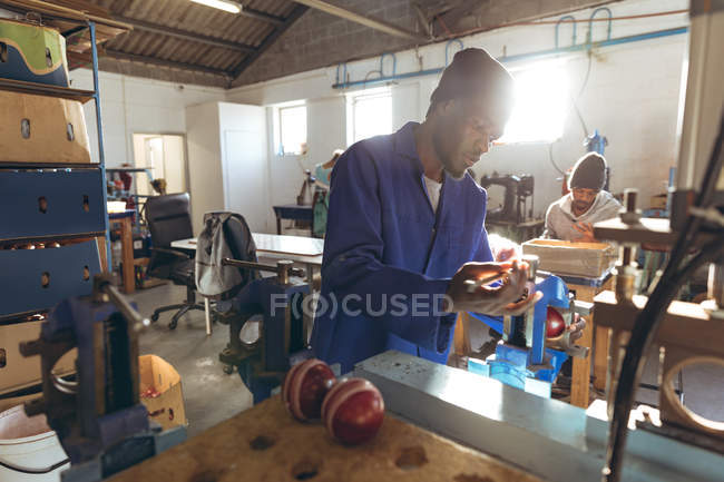 Vista lateral de cerca de un joven afroamericano operando una máquina en un taller en una fábrica de bolas de cricket, en el fondo un colega afroamericano está sentado en un banco de trabajo trabajando en otra parte de la línea de producción . - foto de stock