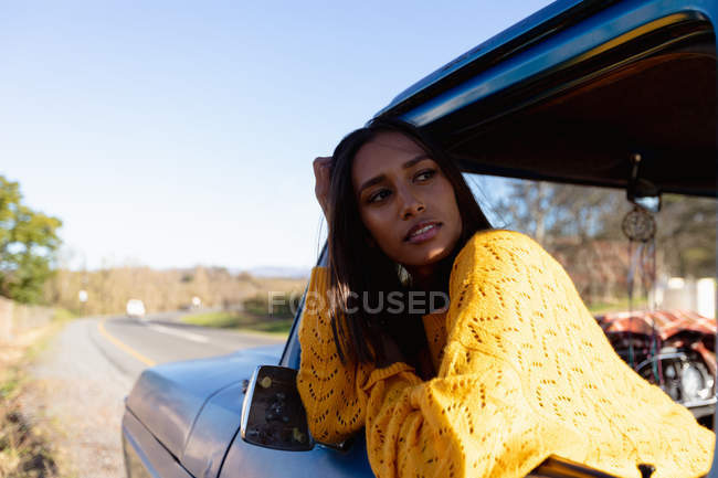 Retrato de una joven mujer de raza mixta sentada en el asiento delantero del pasajero de una camioneta, inclinada por la ventana lateral y mirando a su alrededor durante un viaje por carretera - foto de stock