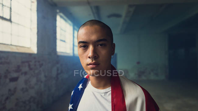 Frontansicht eines jungen hispanisch-amerikanischen Mannes in einem schlichten weißen Hemd mit einer amerikanischen Flagge über den Schultern, der in einer leeren Lagerhalle aufmerksam in die Kamera blickt — Stockfoto