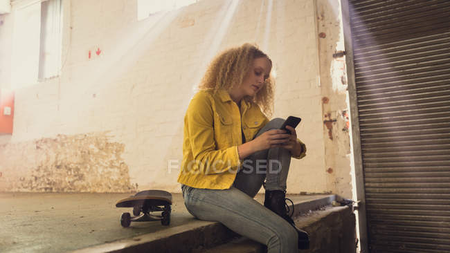 Вид збоку молода кавказька жінка з Кучеряве волосся носити жовту куртку над сірої сорочці сидить поруч з скейтбордом при використанні мобільного телефону всередині порожній склад — стокове фото