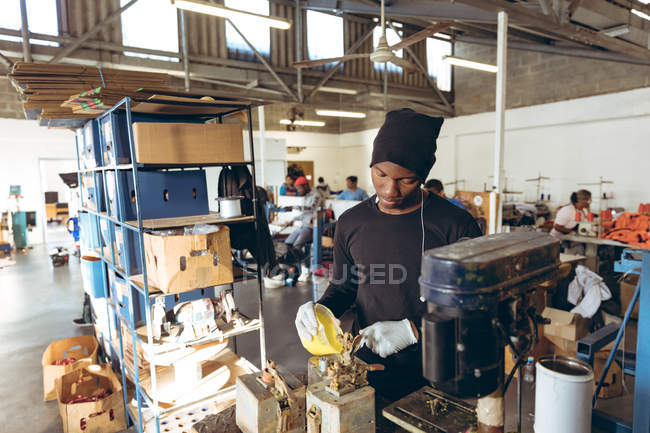 Front view of a young African American man wearing a hat standing at a workbench pouring rubber into a mold in a workshop at a factory making cricket balls, in the background colleagues are working on other parts of the production line. — Stock Photo