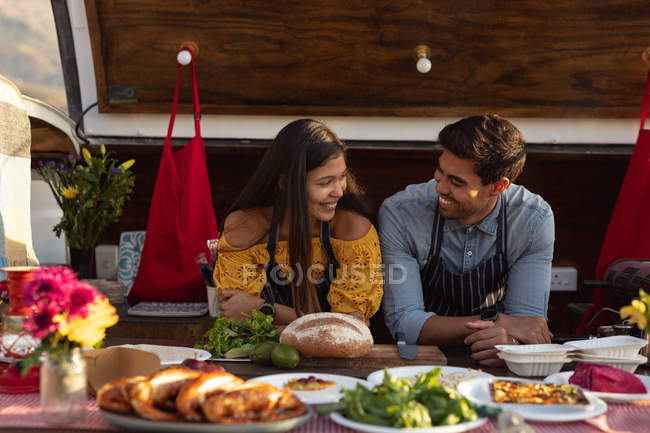 Front view close up of a young mixed race couple smiling at each other sitting in an open top van offering a range of takeaway food for sale — Stock Photo