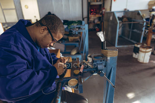 Vista lateral de cerca de un joven afroamericano con gafas de pie en un banco de trabajo sosteniendo el hilo en la boca y las manos, cosiendo una pelota en un taller en una fábrica de bolas de cricket . - foto de stock