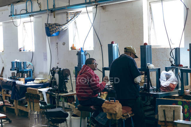 Vista trasera de dos jóvenes trabajadores masculinos de raza mixta sentados, hablando y operando máquinas en una fábrica de bolas de cricket . - foto de stock